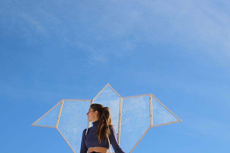 photograpphie d'une fille en contre-plongée avec un objet en forme de deltaplane dans le dos sur un fond de ciel.