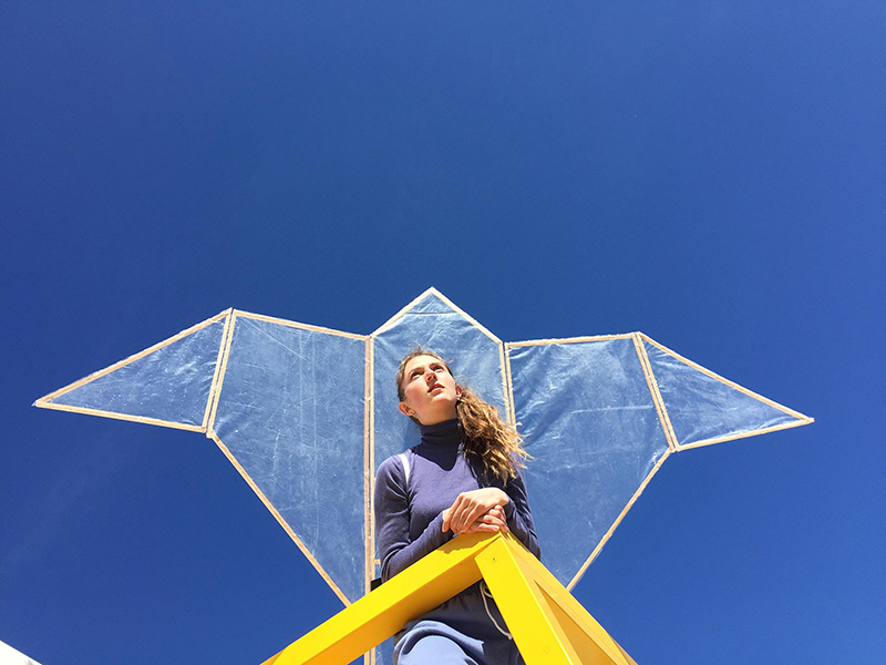 photographie d'une fille en contre-plongée sur une rembarde avec un objet en forme de deltaplane dans le dos sur un fond de ciel.