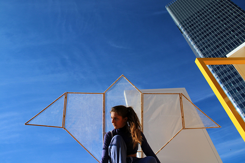 photographie d'une fille en contre-plongée agenouillée avec un objet en forme de deltaplane dans le dos sur un fond de ciel.