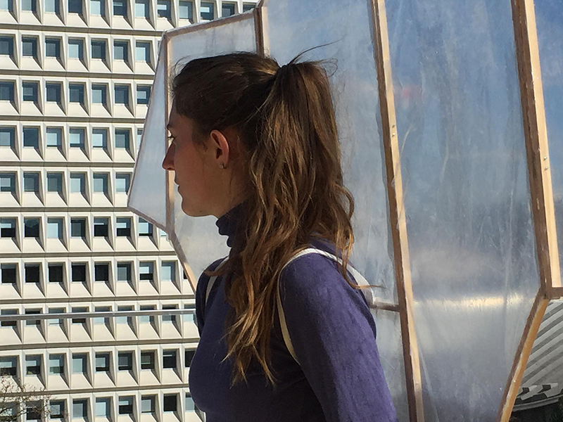 photographie d'une fille de profil avec un objet en forme de deltaplane dans le dos et un bâtiment en arrière plan.