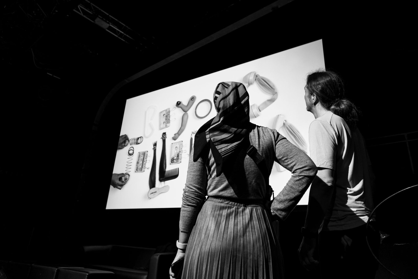 un homme et une femme participant à Beyon Tellerrand de dos devant un écran de projection sur lequel sont affichés des objets relatifs à une conférence.